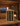 Wine rack with bottles, a candle, and a glass of wine on a wooden table in a cozy room with a fireplace.