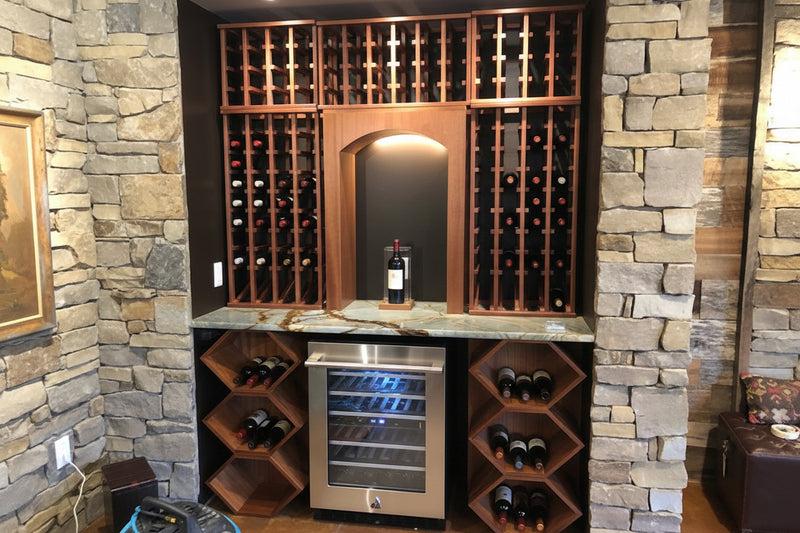 Wine cellar with wooden racks and a stainless steel wine cooler against a stone wall.