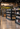 A wide, well-lit wine store aisle with multiple black shelving units stocked with wine bottles. The space has polished wood floors, hanging pendant lights, and large landscape wall murals in the background.