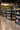A wide, well-lit wine store aisle with multiple black shelving units stocked with wine bottles. The space has polished wood floors, hanging pendant lights, and large landscape wall murals in the background.
